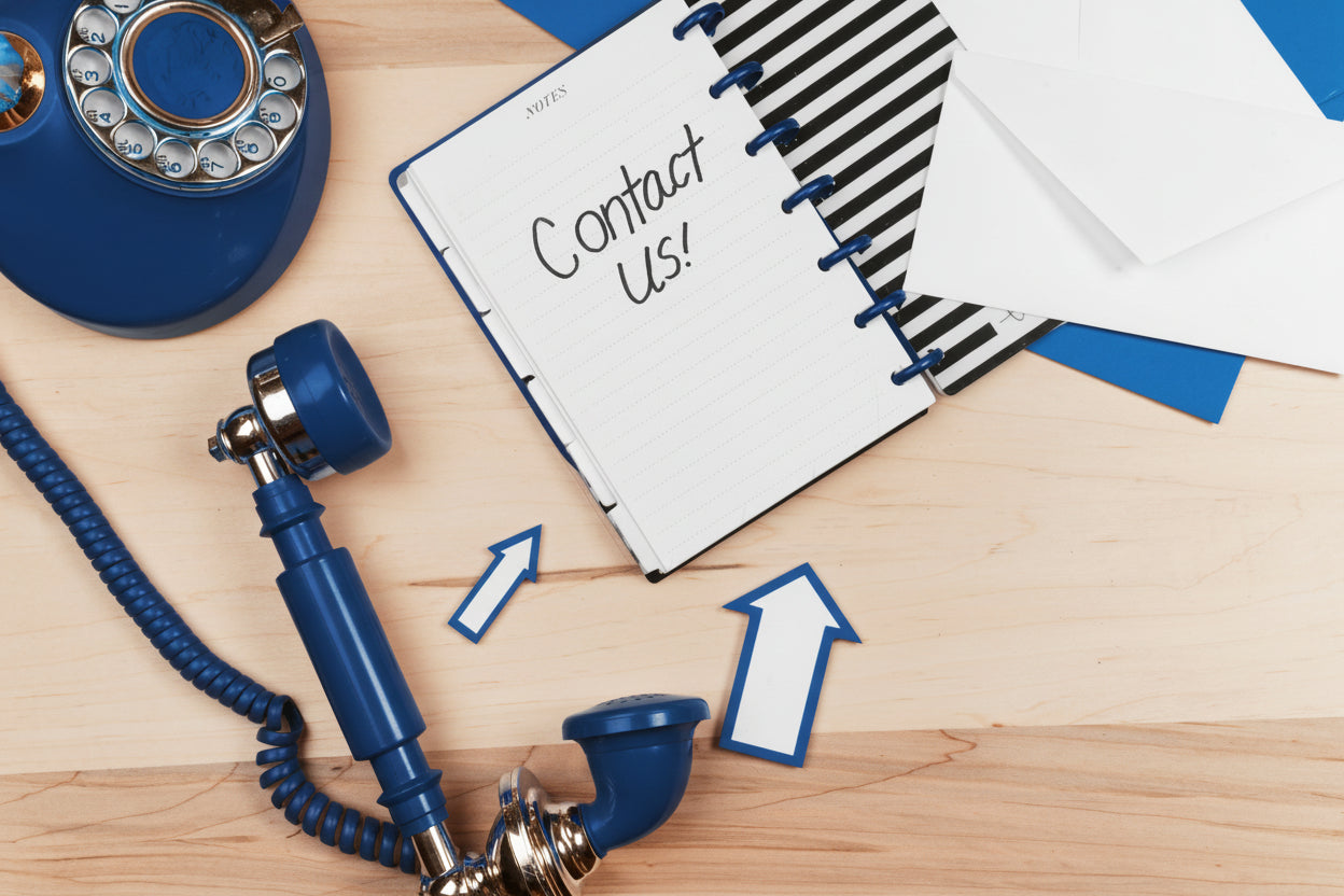 A flat-lay view of a vintage blue rotary phone next to a notebook with "Contact Us!" handwritten on a page, accompanied by white envelopes on a light wooden desk surface.