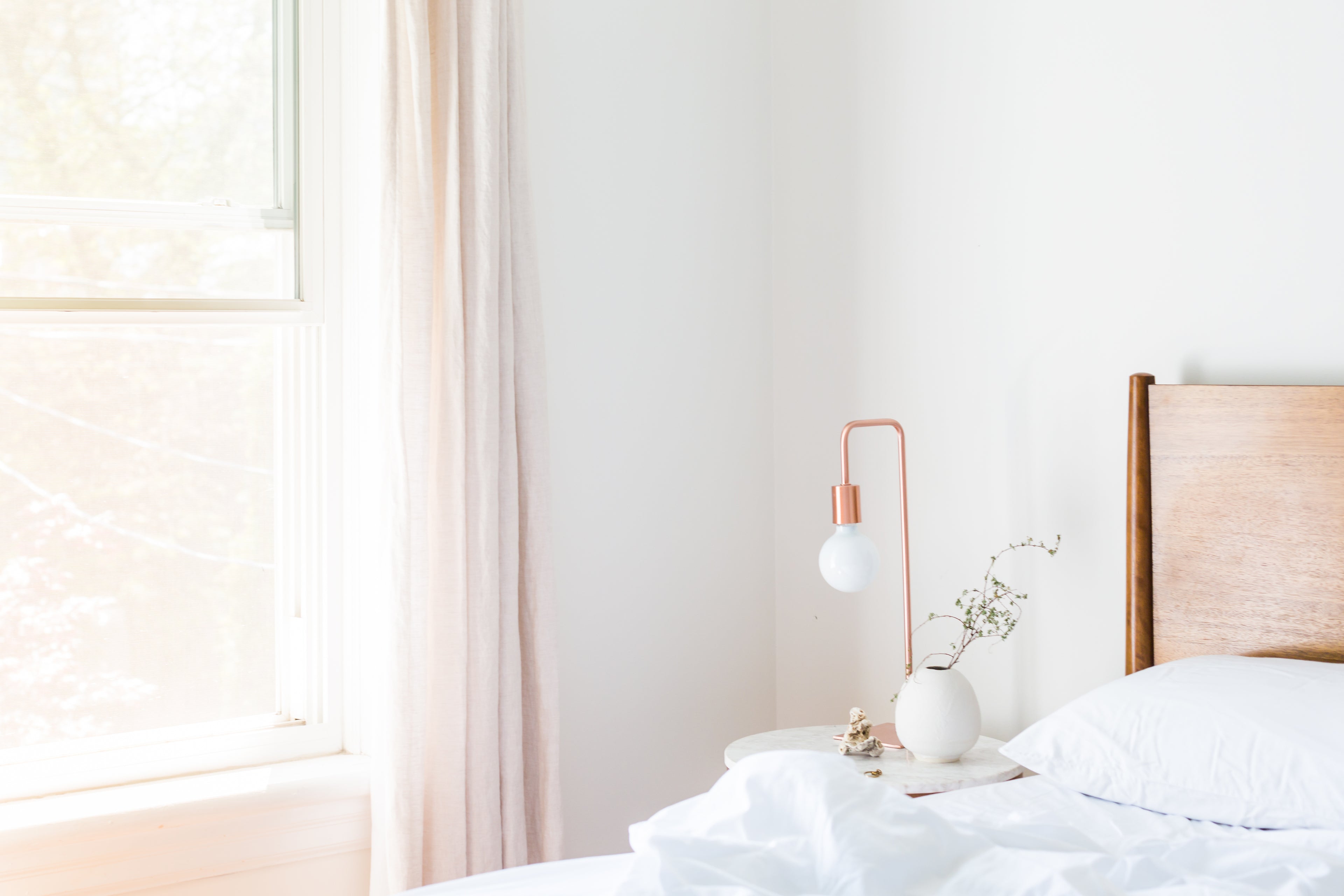 Homellence Modern Bedroom Decor - Minimalist bedroom featuring a white marble bedside table with a copper lamp, a small white vase with greenery, and a wooden headboard in a bright, airy setting.